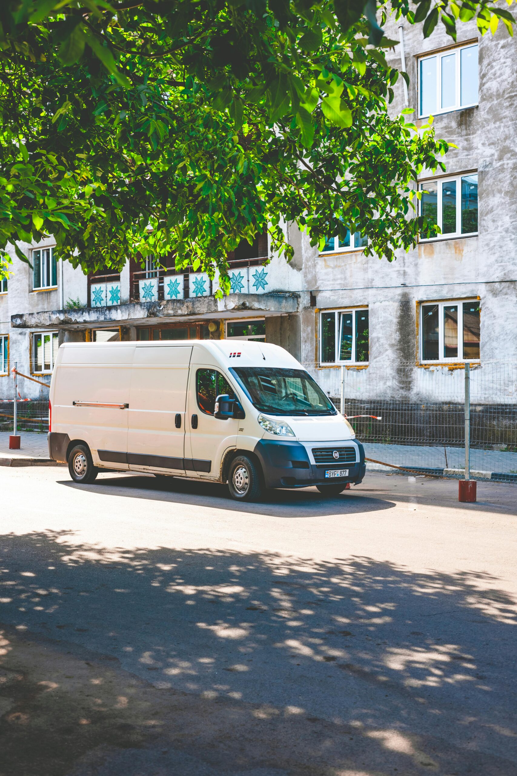 A white delivery van parked under a tree on a sunny day in an urban area.